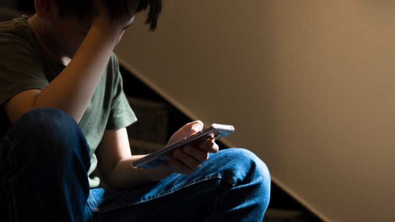 Ein Kind sitzt auf der Treppe, in der einen Hand h&auml;lt er ein Smartphone, mit der anderen st&uuml;tzt er seinen Kopf